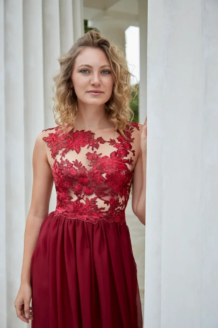 Model in einem eleganten roten Kleid mit Spitzenoberteil, stehend vor weißen Säulen.