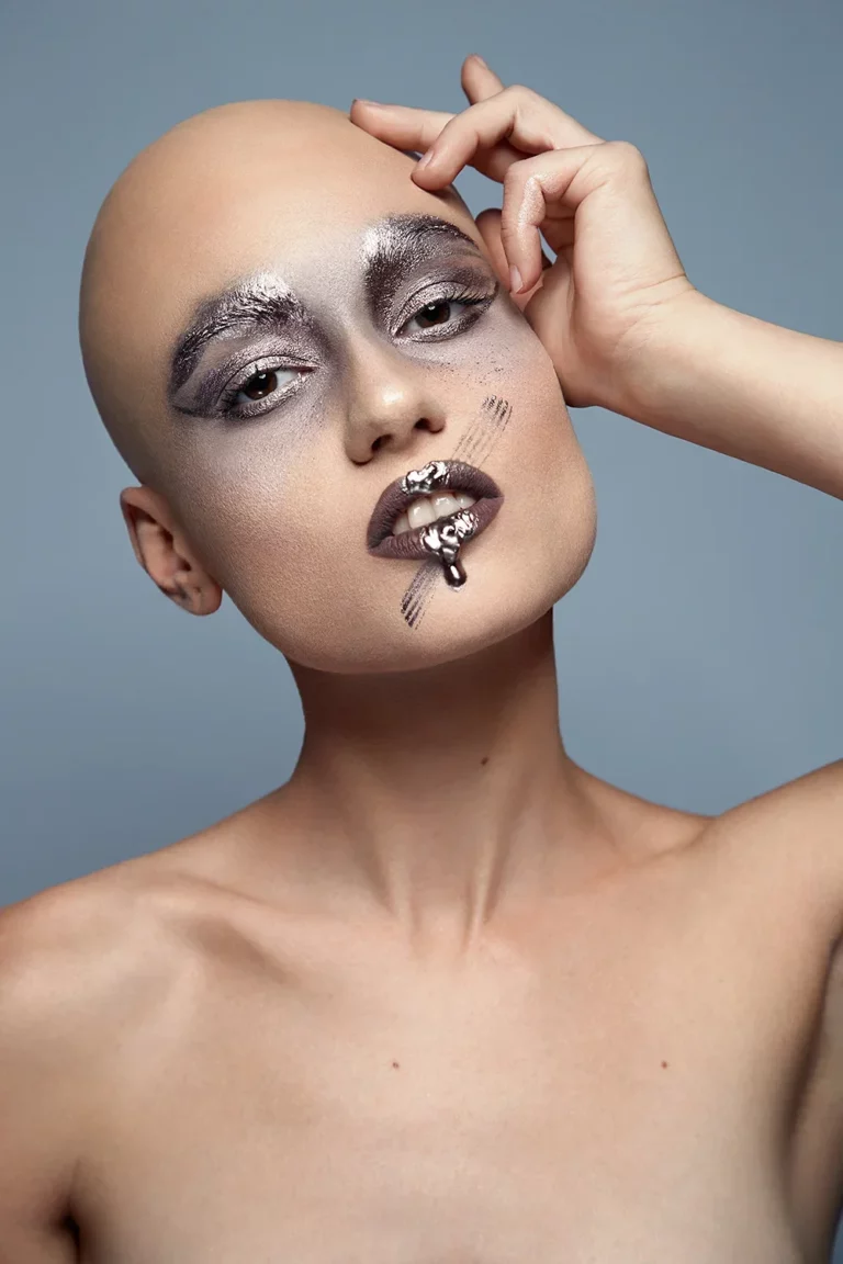 Glatzköpfige Frau mit futuristischem silbernem Make-up und metallischem Lippenstift, eine Hand an der Schläfe, vor einem hellblauen Hintergrund.