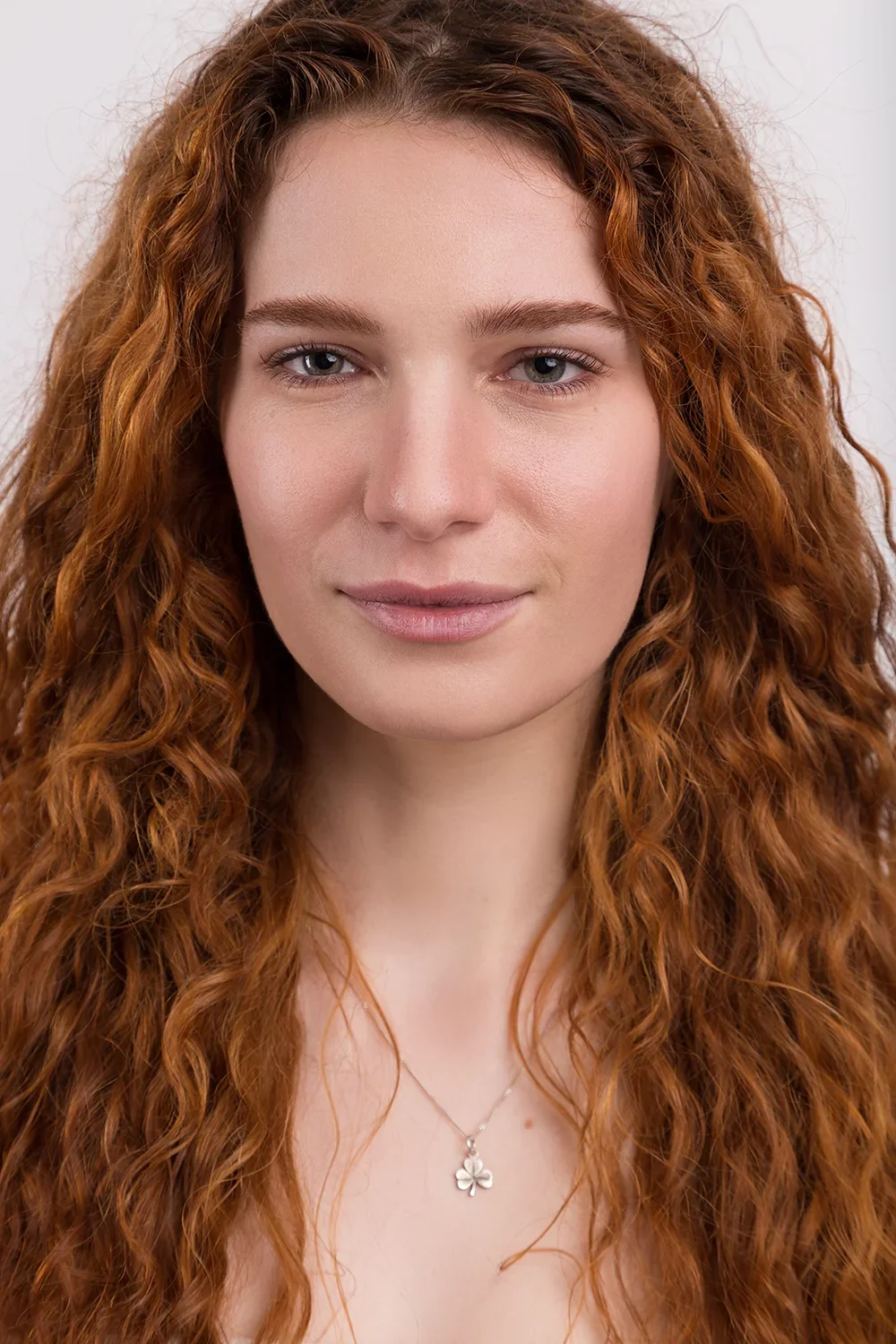 Model mit langen, lockigen rotbraunen Haaren und einem Kleeblatt-Anhänger, vor einem weißen Hintergrund.