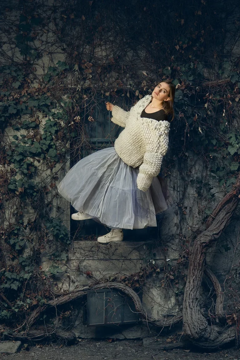 Model in voluminösem Pullover und blauem Tüllrock steht am Fenstersims und hält sich am Fenstergitter fest. Umgeben ist sie von Efeu.