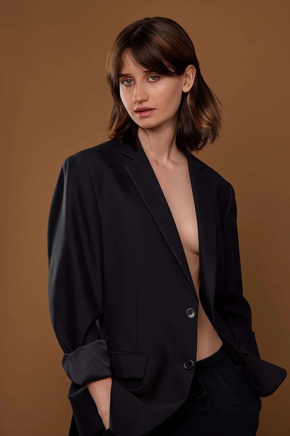 Model in offenem schwarzen Blazer ohne Hemd vor braunem Hintergrund.
